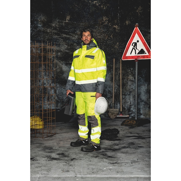 Person in gelber Warnschutzjacke und -hose mit Reflexstreifen, Helm in der Hand, neben einem Baustellenschild.